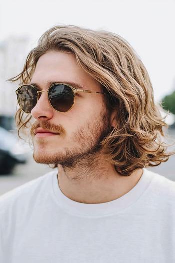 A man with medium-length wavy hair, wearing round sunglasses and a white shirt, with facial hair.