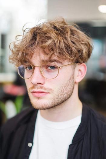 A young man with tousled, medium-length, wavy hair and wearing glasses.