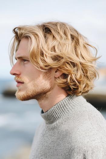 A man with medium length, wavy blonde hair wearing a grey turtleneck sweater.