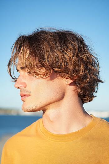 A man with wavy, medium-length hair, styled in loose, natural waves, wearing a yellow shirt and looking sideways.