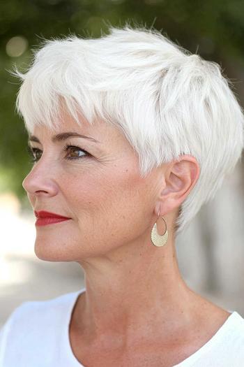 A woman with short, white, textured hair in a pixie cut, wearing red lipstick and gold earrings.