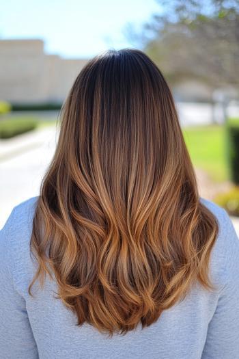 A person with long, wavy hair featuring caramel balayage, blending dark brown roots with warm caramel highlights towards the ends.