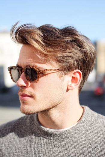 The image shows a man with a medium length hairstyle featuring swept-back hair and a slight side part, paired with sunglasses and casual attire.